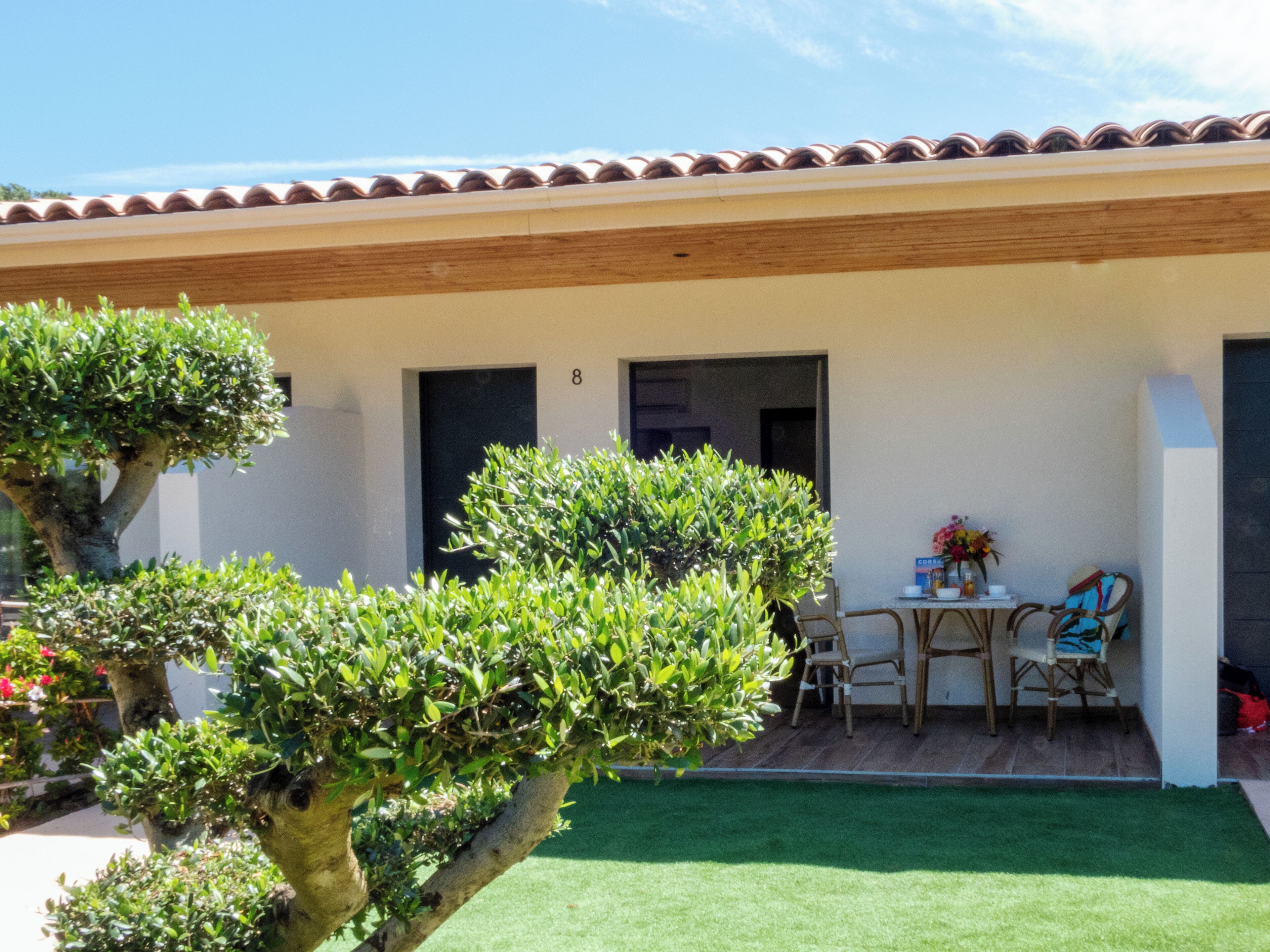 A covered patio with a table and chairs, a small garden, and a building with a tiled roof.