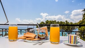 A table with two glasses of orange juice, a plate with bread and a jar of jam, and a striped mug, set against a scenic view of the sea and trees. - Theodora Apartments (West Mani)