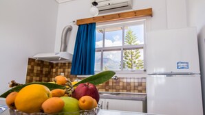 A kitchen with a fruit bowl on the counter, a window with a view of trees, and a white refrigerator. - Theodora Apartments (West Mani)