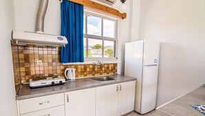 A kitchen with white cabinets, a stove, a kettle, a sink, and a window with blue curtains. - Theodora Apartments (West Mani)