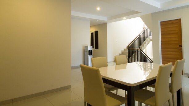 A modern dining area with a white table and four chairs, a staircase with a wooden door, and a refrigerator in the background. - RedDoorz Plus @ Setiabudi Tengah (Jakarta)
