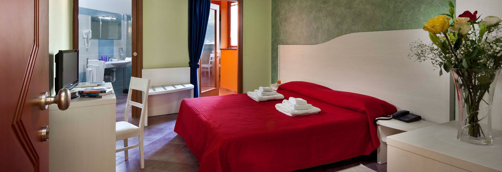 Ein Hotelzimmer mit roter Bettdecke, einem Nachttisch mit Telefon, einer Vase mit Blumen und Blick auf das Badezimmer und den Balkon.