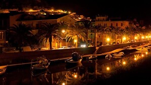 Vista nocturna de una zona costera con barcos amarrados, palmeras bordeando el paseo marítimo y edificios con iluminación cálida al fondo.
