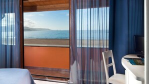 Un dormitorio con cama, una silla, una mesita y televisor. La habitación tiene vista a la playa y al mar.