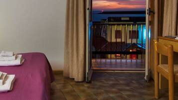 A hotel room with a bed, a desk, and a balcony with a view of the sunset.