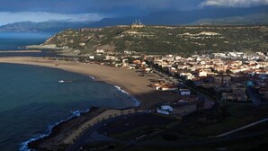 Vista aérea de un pueblo costero con una fortificación destacada, una playa y un paisaje montañoso.