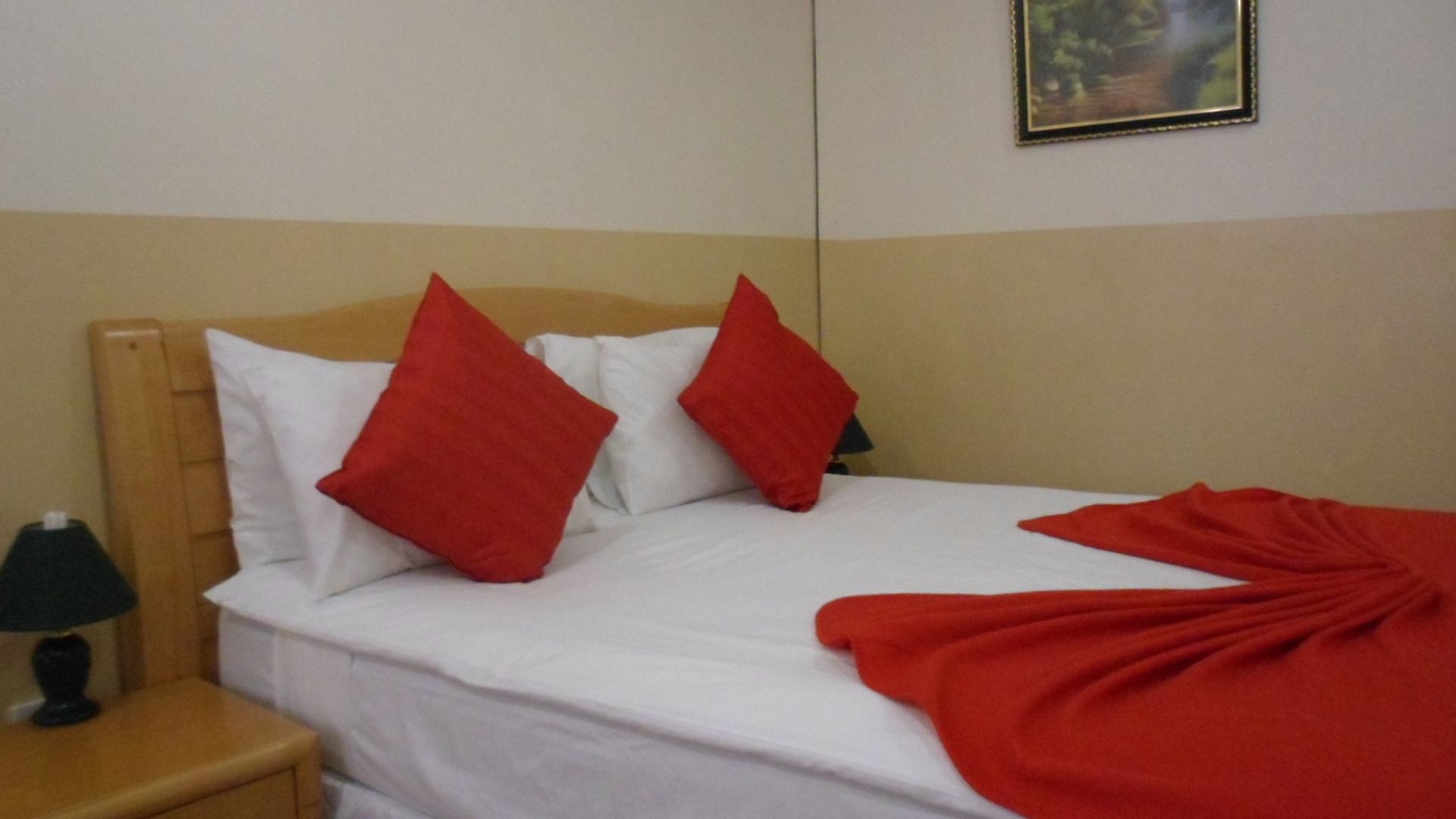 A bed with white bedding and two red pillows, a wooden headboard, a nightstand with a lamp, and a red blanket on the bed.