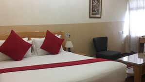 Hotellihuone, jossa on sänky, punaiset ja valkoiset tyynyt, puinen pääty, pieni pöytä, tuoli ja taulu seinällä.
