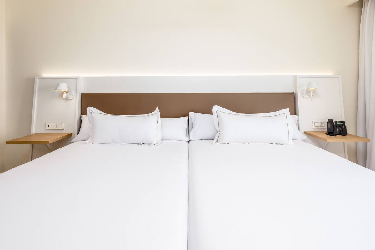 Un lit double avec du linge de lit blanc, deux tables de chevet et des lampes fixées au mur.