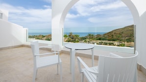 Een balkon met witte stoelen en een tafel, met uitzicht op een kustlandschap met heuvels en palmbomen.