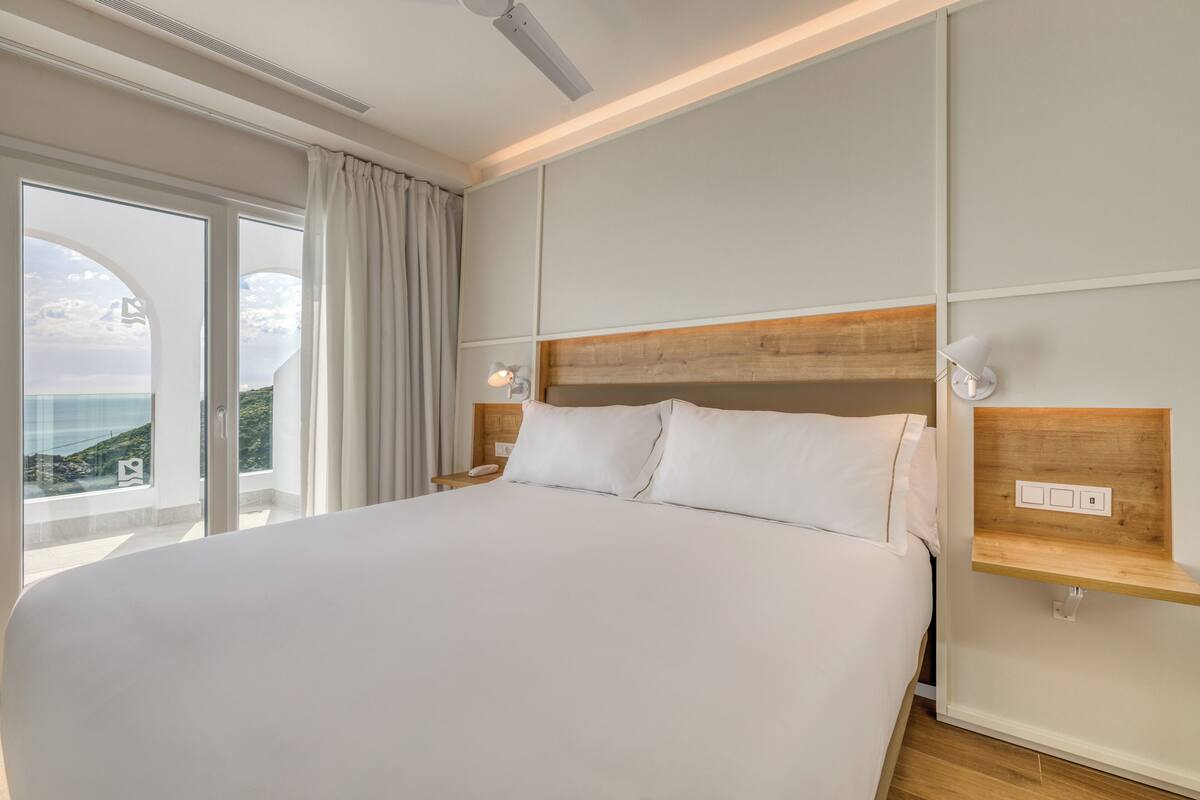 Un grand lit avec du linge de lit blanc, une tête de lit en bois, une table de chevet avec une lampe, et une vue sur la mer par une grande fenêtre avec des rideaux.
