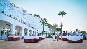 Een hotel met een witte gevel, een terras met tafels en palmbomen.