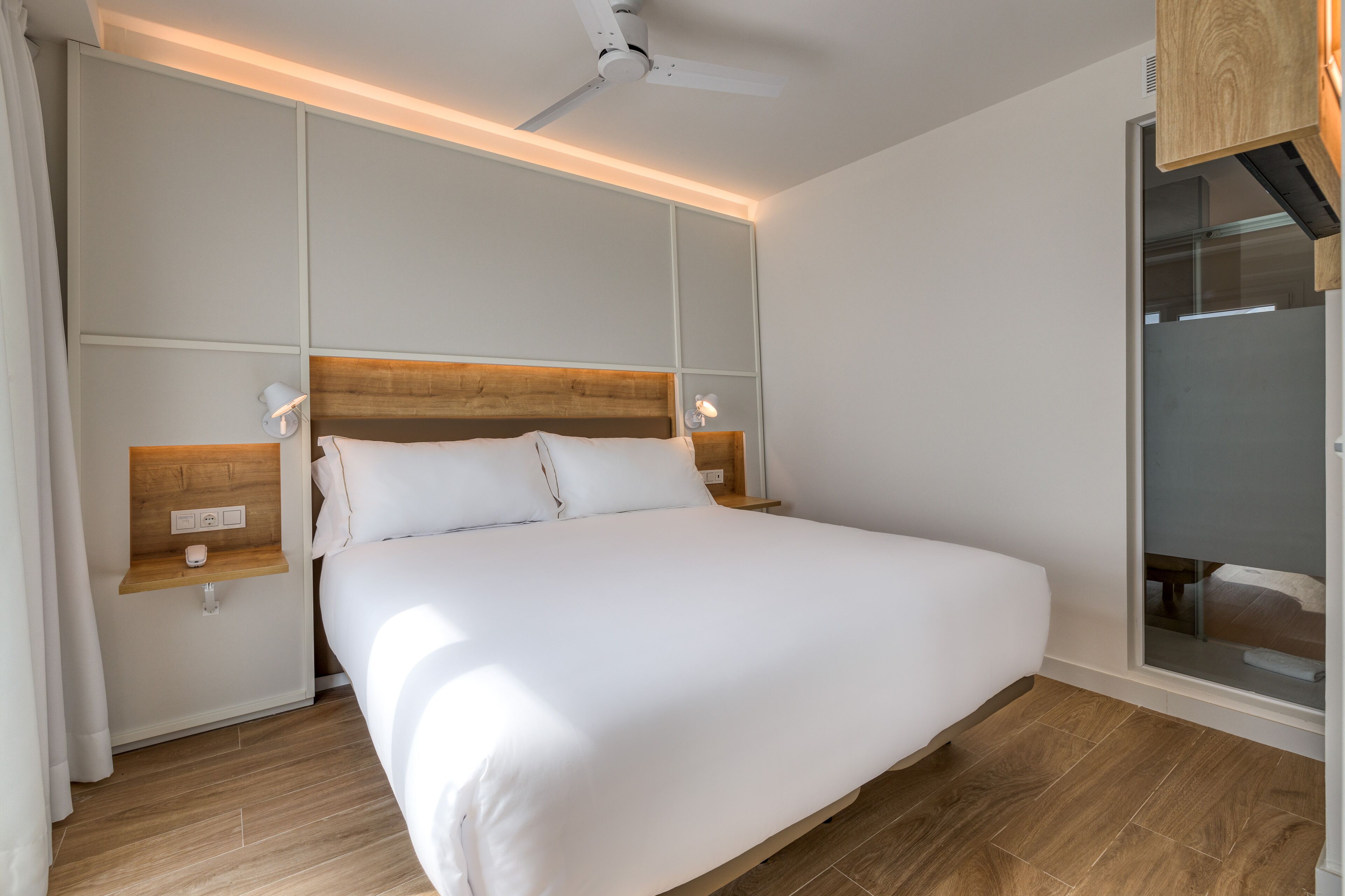 A modern hotel room with a large bed, wooden headboard, bedside tables with lamps, and a ceiling fan.