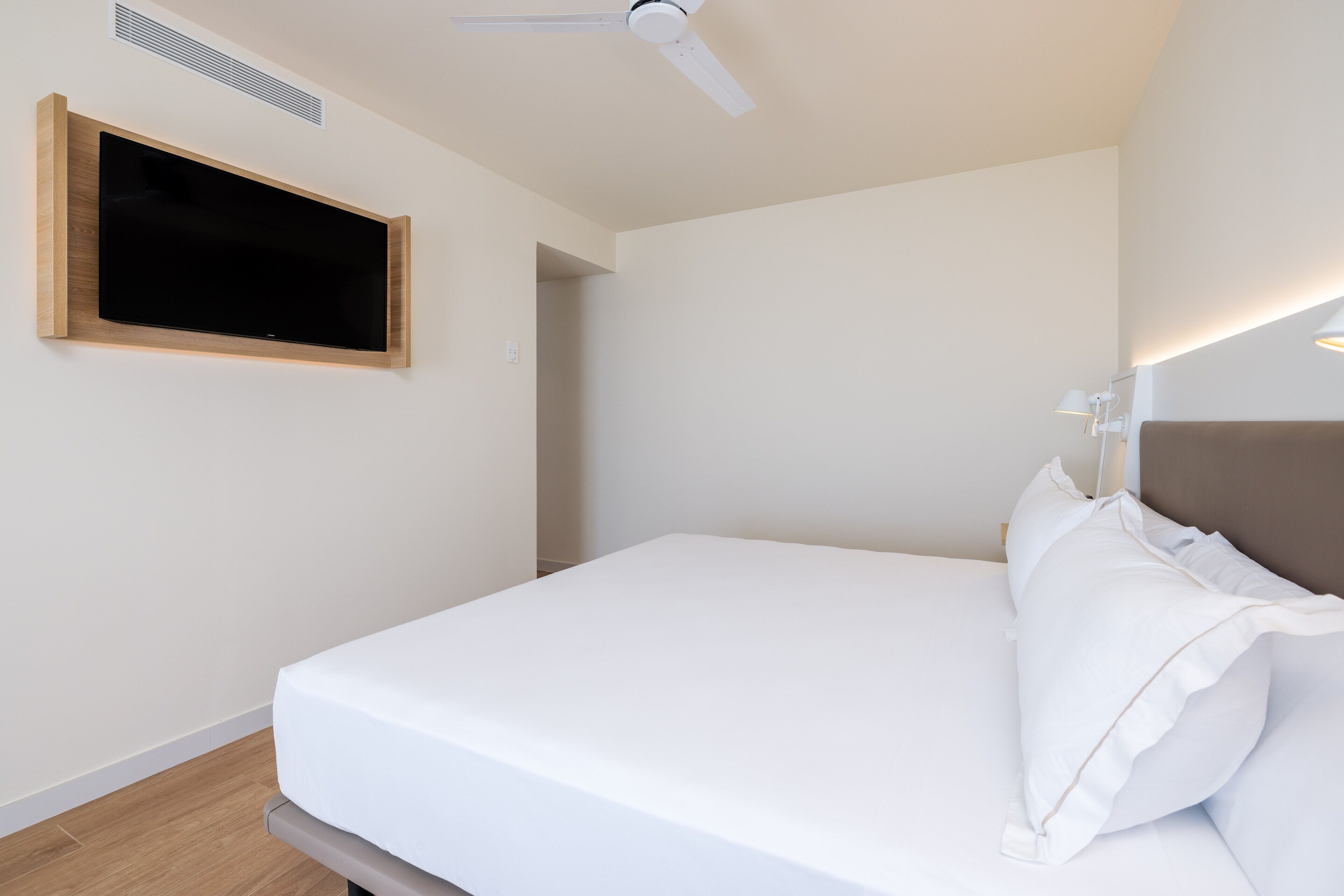 A bedroom with a bed, a ceiling fan, a mounted TV, and a bedside lamp.