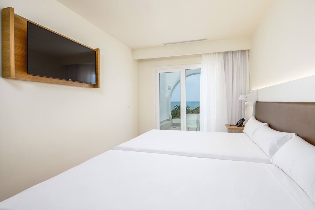 Une chambre d’hôtel dotée d’un grand lit, d’un téléviseur à écran plat fixé au mur, d’une table de chevet avec une lampe et offrant une vue sur l’extérieur grâce à une porte-fenêtre coulissante.