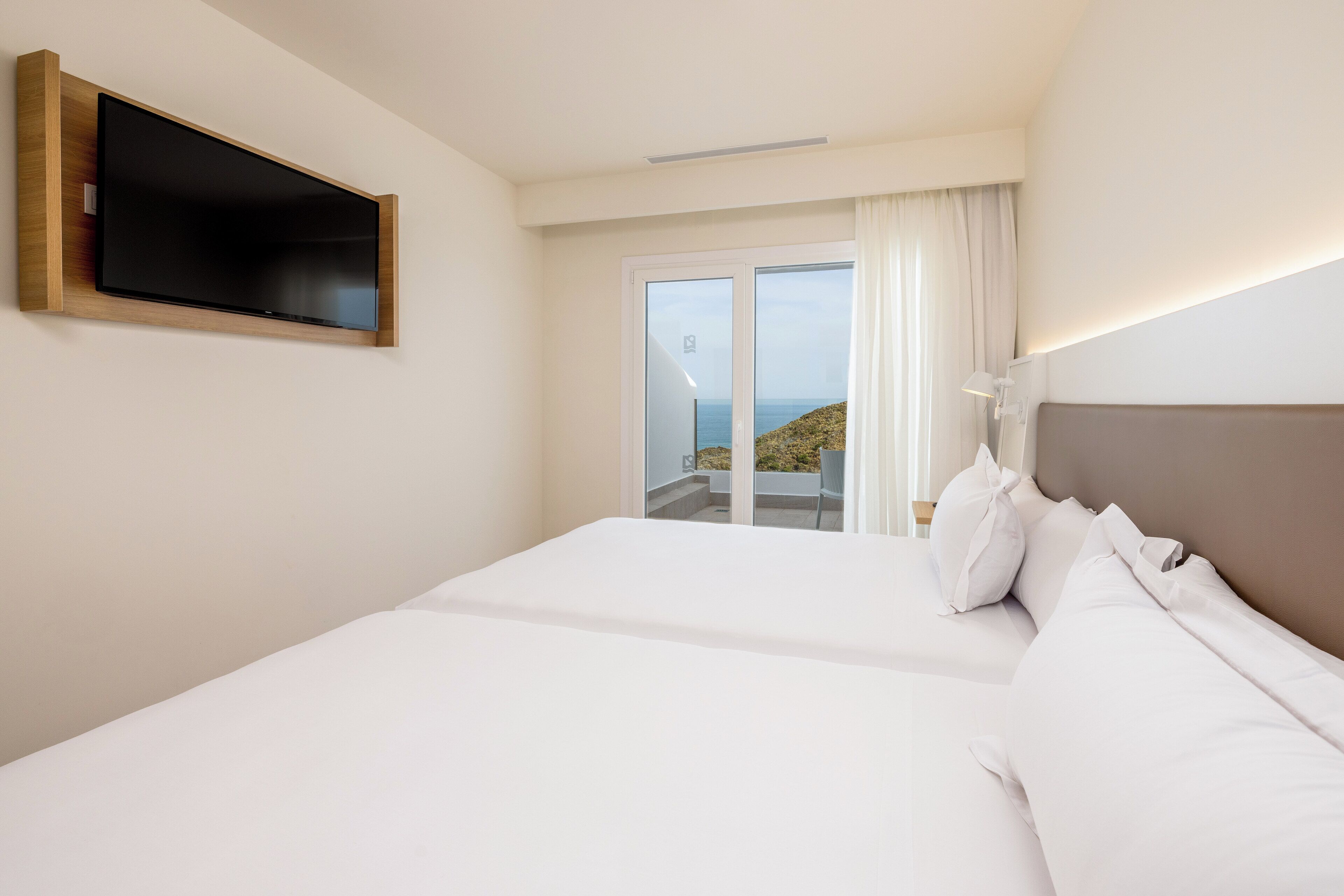 A hotel room with a large bed, a flat-screen TV mounted on the wall, and a view of the sea through a sliding glass door.