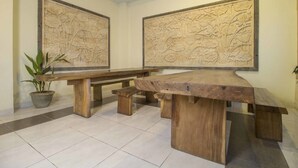 A spacious room with a large wooden table and benches, two large wall carvings, and a potted plant. - RedDoorz @ Benesari 2 (Kuta)
