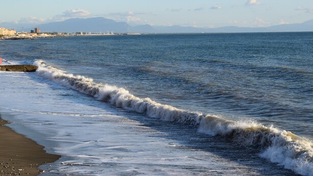 A coastal scene with waves approaching the shore, a clear sky, and distant mountains.