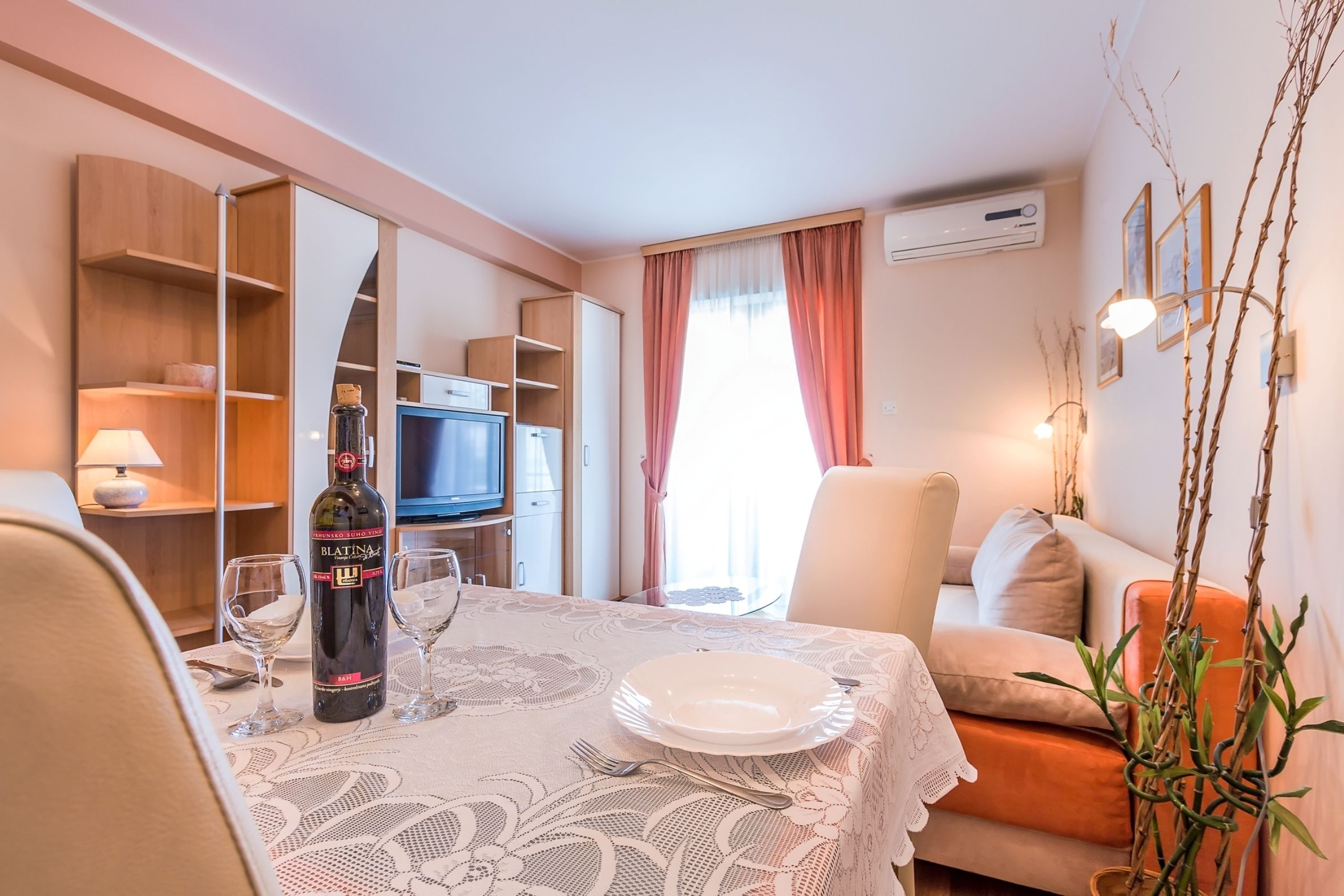 Un coin repas avec une table dressée pour deux personnes, une bouteille de vin Blatinus et un téléviseur en arrière-plan.