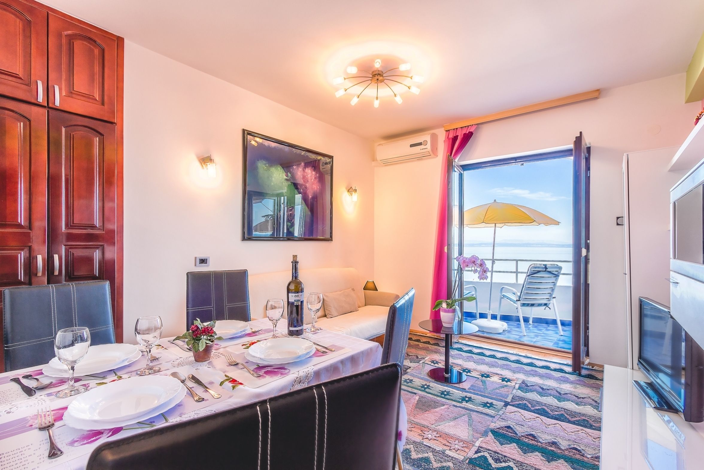 Uma sala de jantar com mesa posta para quatro pessoas, um sofá e uma varanda com um guarda-sol amarelo.