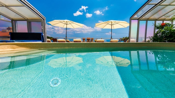 Ein Schwimmbecken mit klarem, blauen Wasser, umgeben von Liegestühlen und Sonnenschirmen, und ein gläsernes Gebäude mit Blick auf den Himmel und die Wolken.