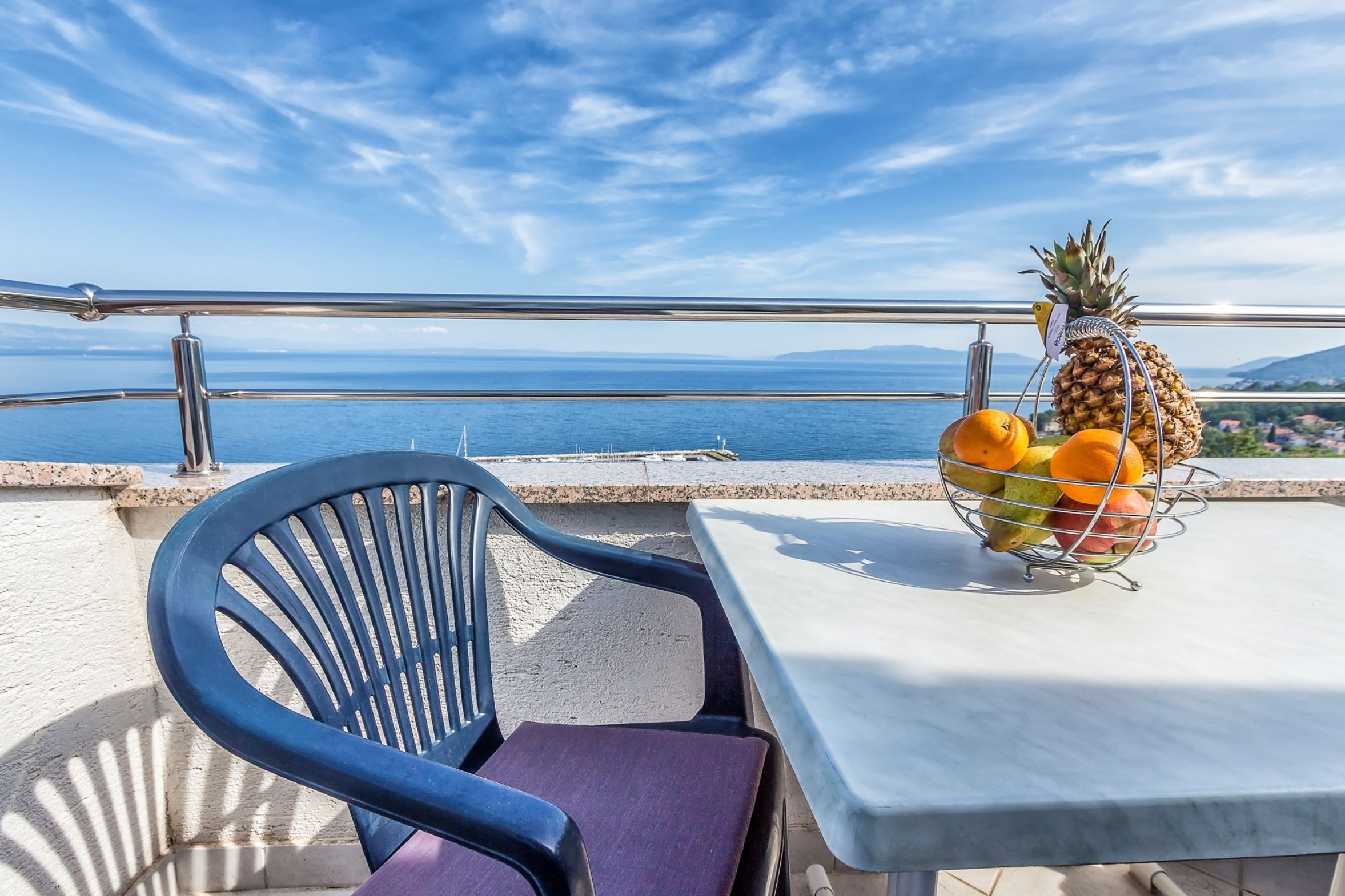 Een balkon met een tafel en stoel, een fruitmand met ananas en sinaasappels, en uitzicht op zee.