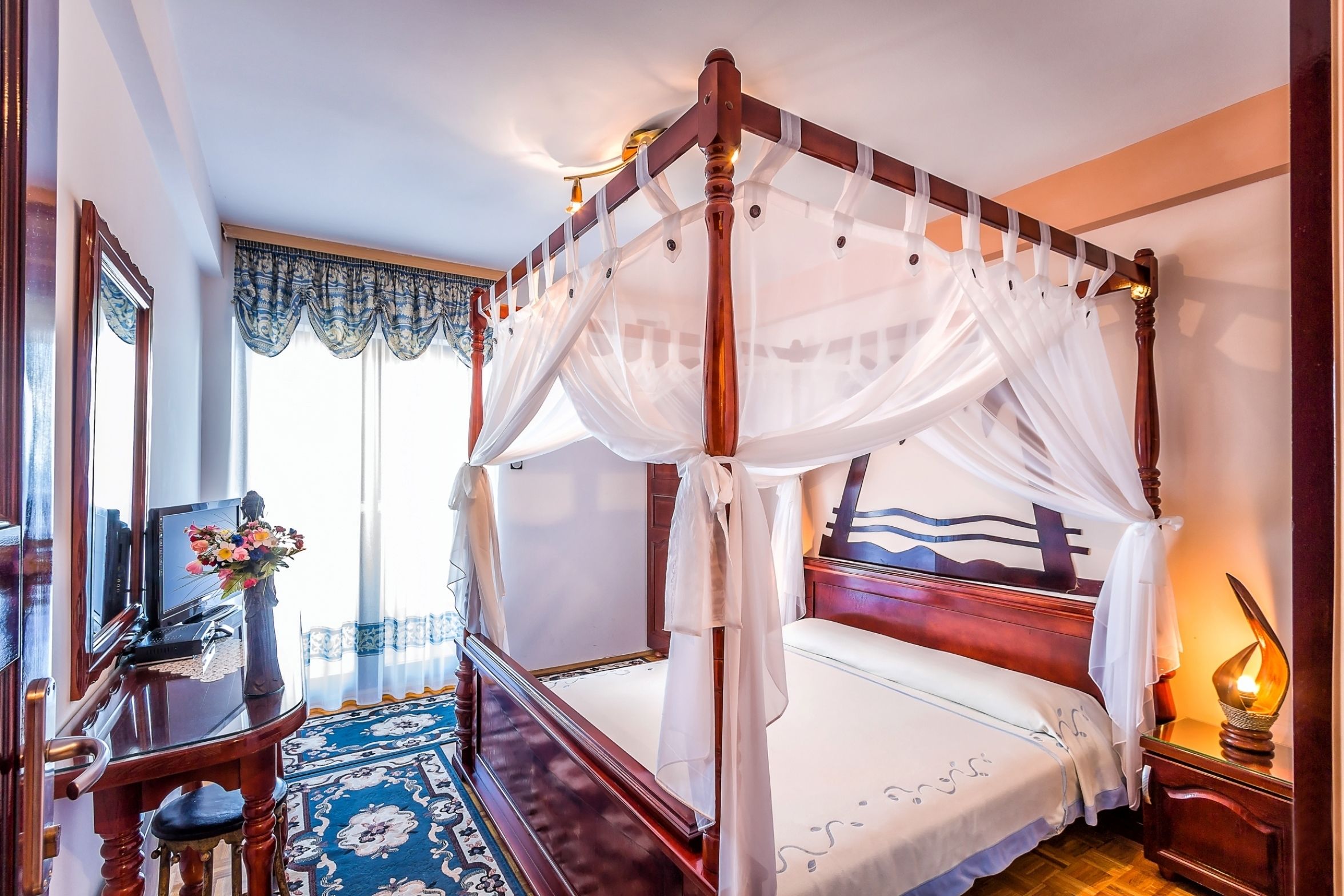 A four-poster bed with a canopy, a bedside table with a lamp, and a decorative vase with flowers.