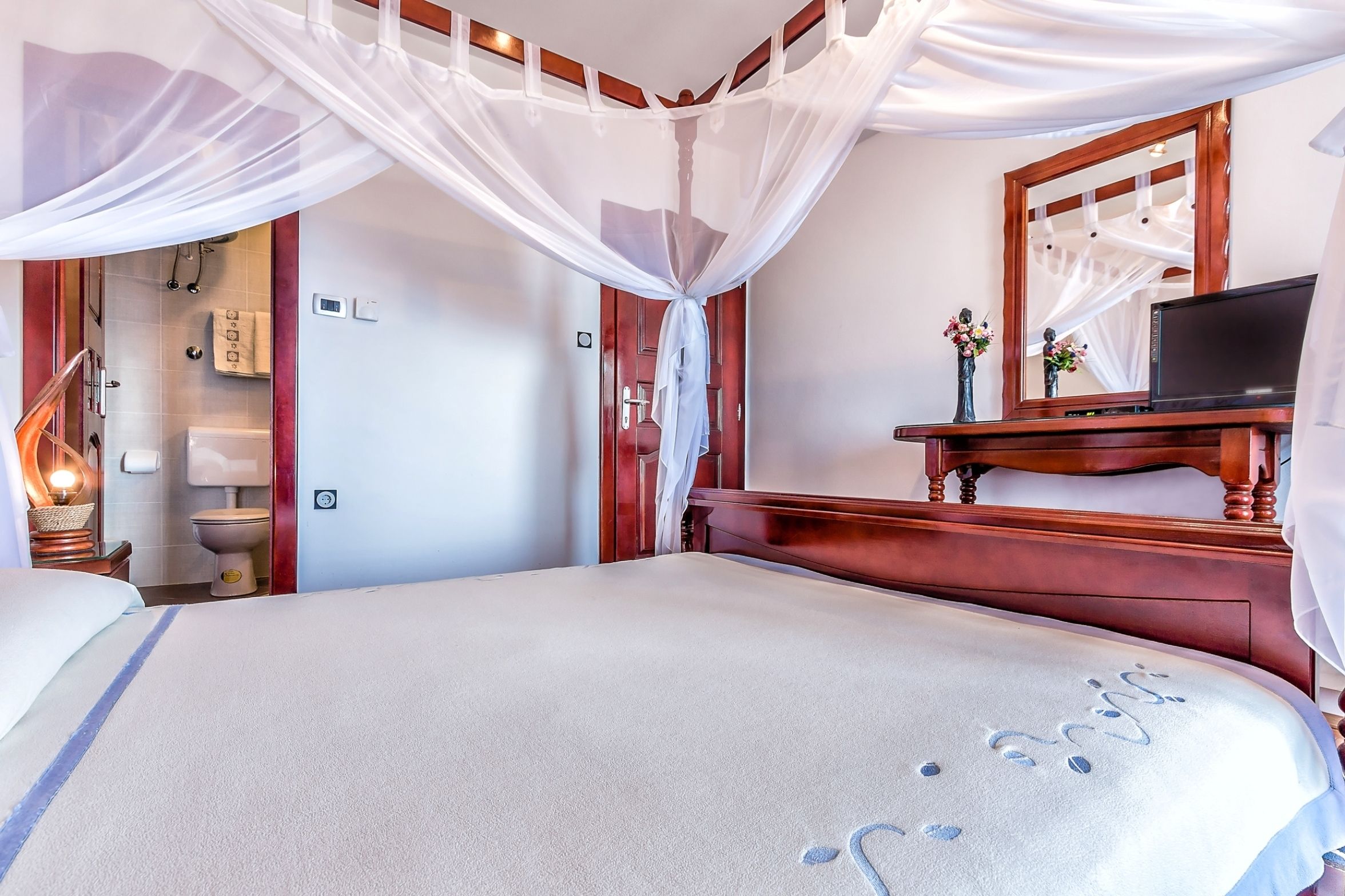 Une chambre d’hôtel avec un lit, une télévision, une tête de lit en bois, un miroir et une salle de bain visible à travers une porte ouverte.