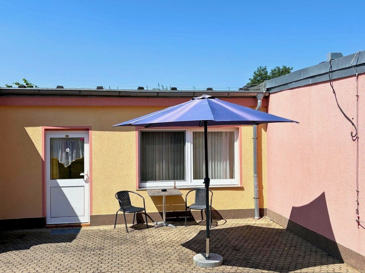 Un pequeño espacio al aire libre con una mesa y dos sillas, una puerta y un ventanal con cortinas.