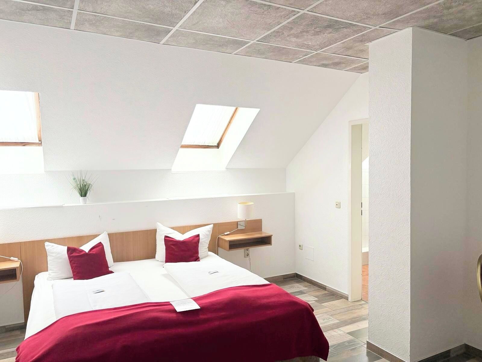 A modern hotel room with a skylight, a bed with white and red bedding, a bedside table with a lamp, and a small plant.