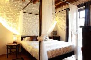 Un dormitorio con pared de piedra, una cama con ropa de cama blanca, una mesita de noche con lámpara y cortinas translúcidas.