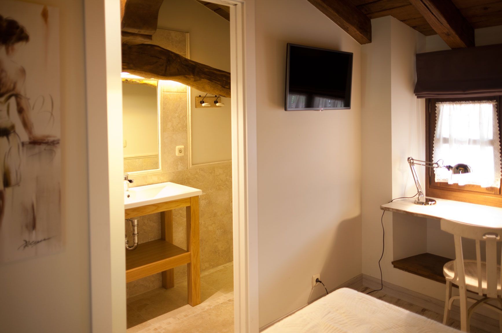 A small, cozy room with a wooden bench, a white desk with a lamp, a TV mounted on the wall, and a window with curtains.