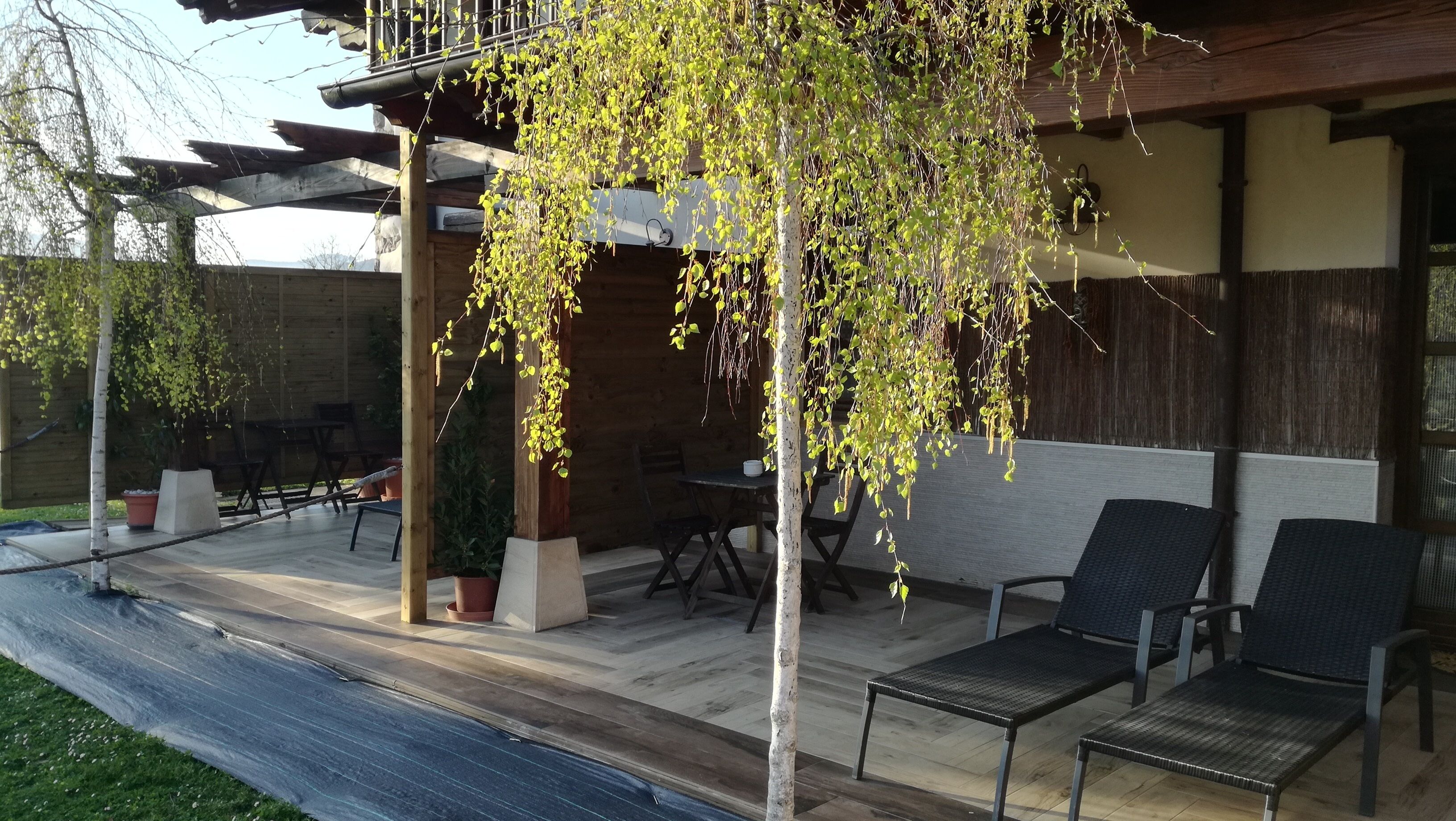 A covered outdoor patio with wooden flooring, a pergola, and a seating area with chairs.