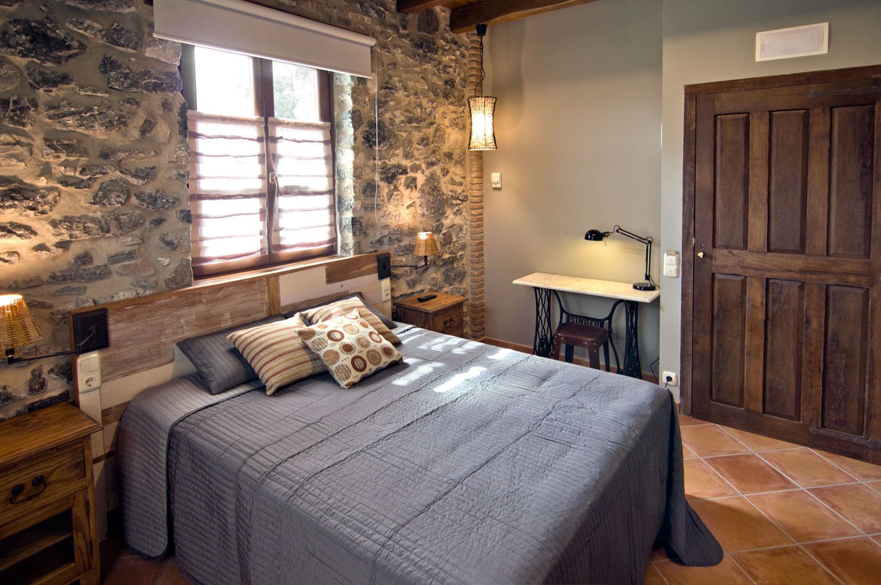 A bedroom with a stone wall, a large bed with a grey quilt, a wooden nightstand, a bedside lamp, and a small table with a lamp.