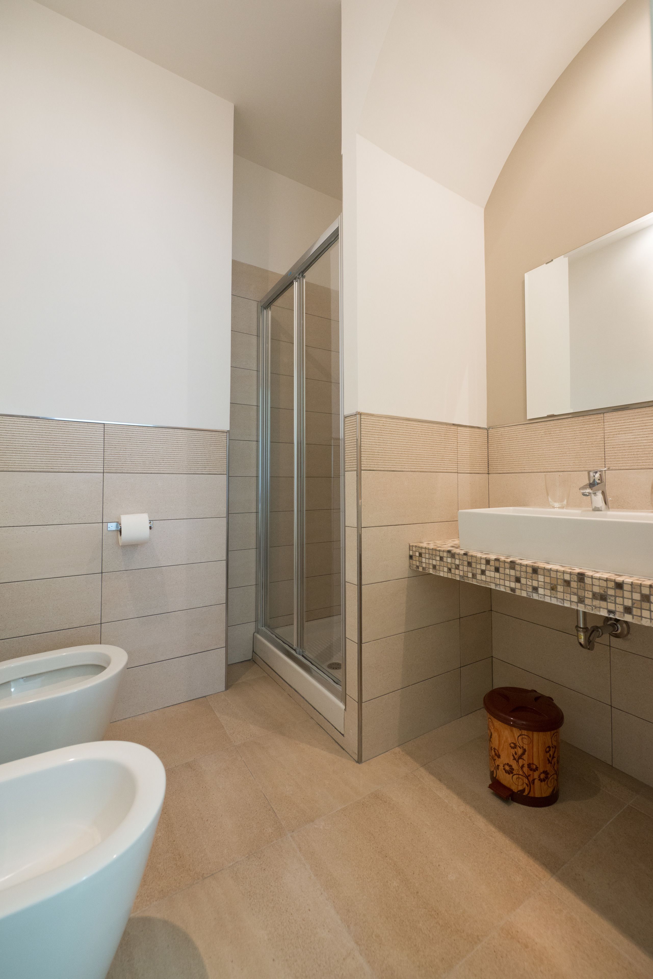 A bathroom with a shower, toilet, bidet, sink, and a trash bin.