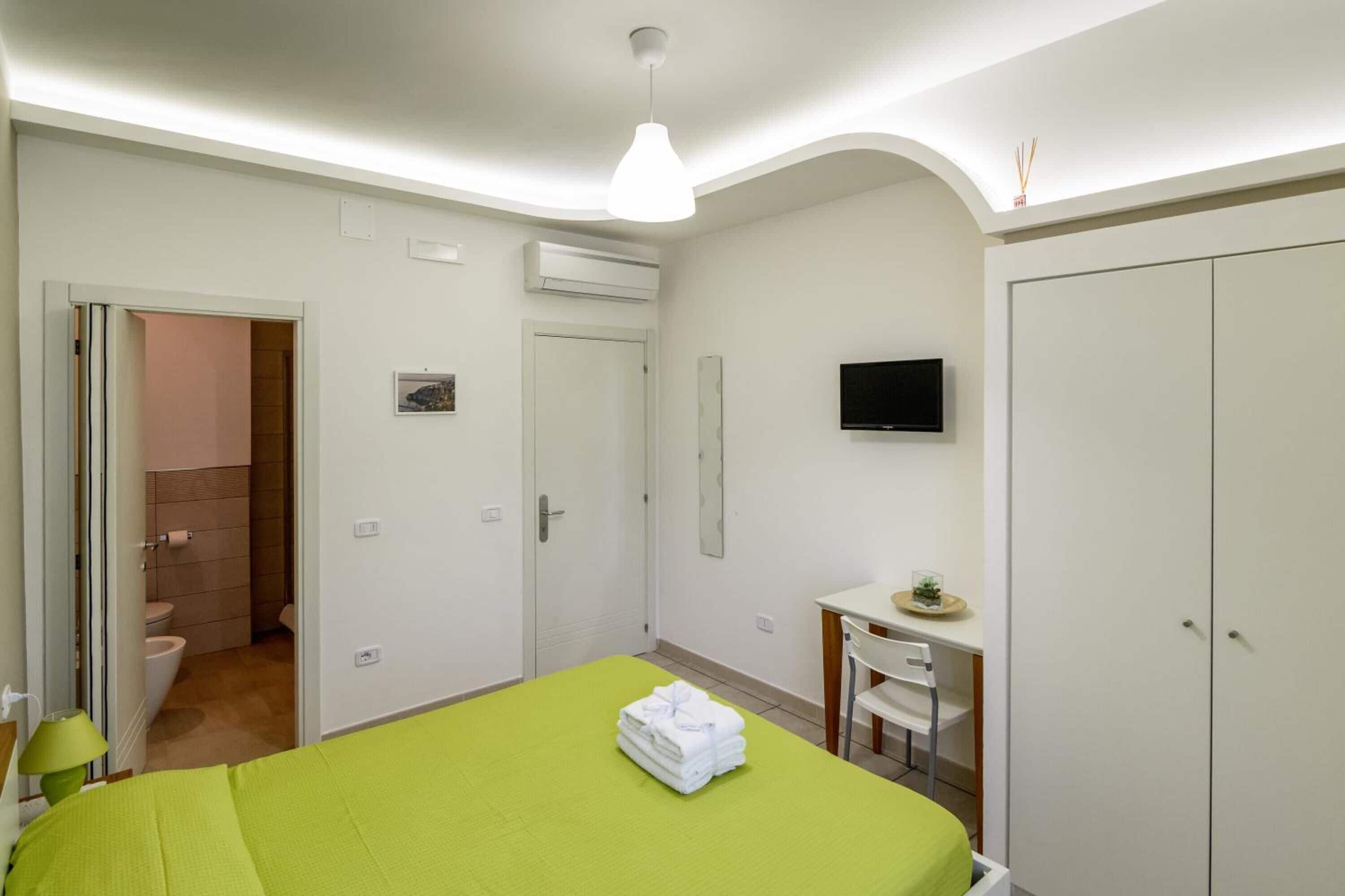 A hotel room with a green bed, a desk with a chair, a TV mounted on the wall, and a wardrobe.