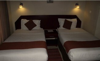 Una habitación doble con dos camas, cada una con ropa de cama blanca y almohadas marrones. Hay mesitas de noche con lámparas y un pequeño estante entre las camas.