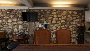Ein Zimmer mit einer Steinwand, einem daran befestigten Fernseher, zwei Holzstühlen, einem kleinen Tisch und einem Bett mit gestreifter Bettdecke.