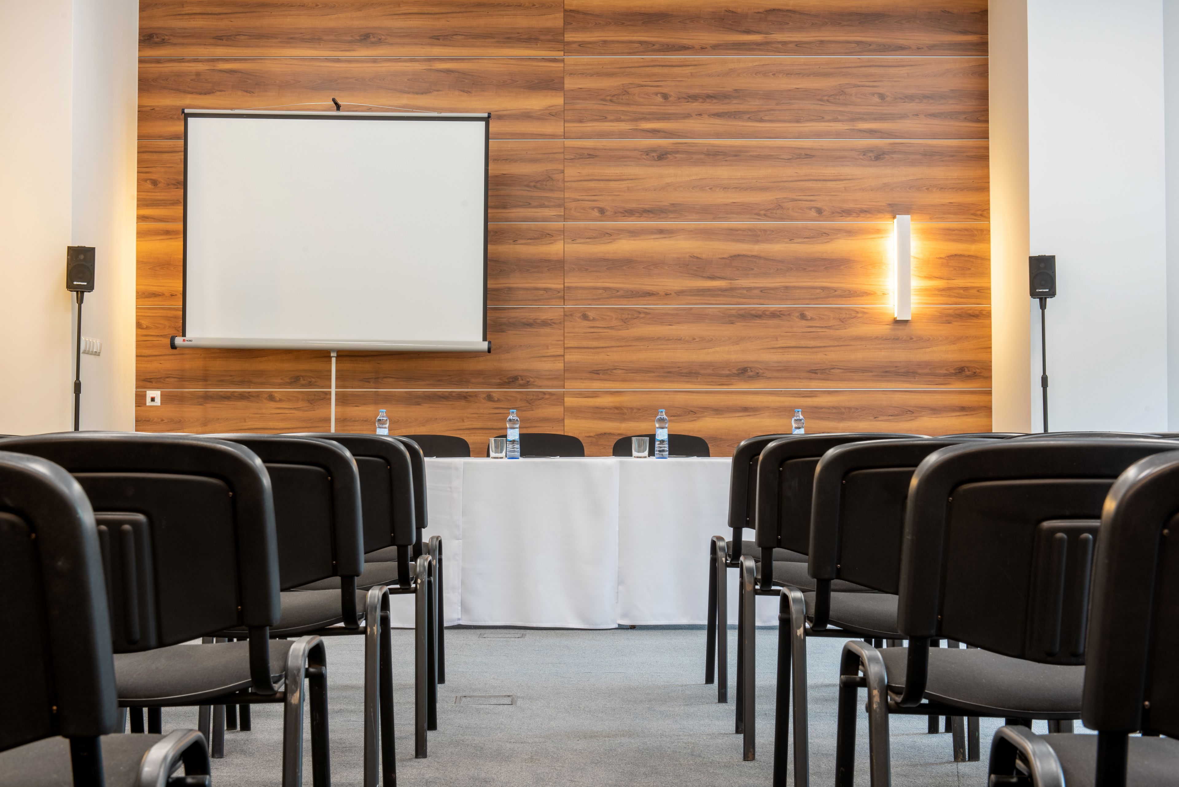 Un salón de conferencias con una pizarra, pared de paneles de madera y filas de sillas negras.