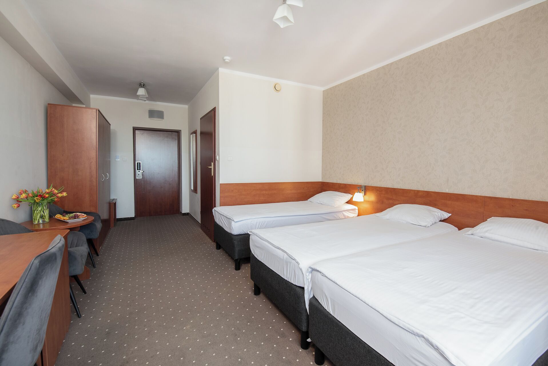 Une chambre d’hôtel avec deux lits simples, un bureau avec une chaise, une armoire et une petite table sur laquelle est posé un vase de fleurs.