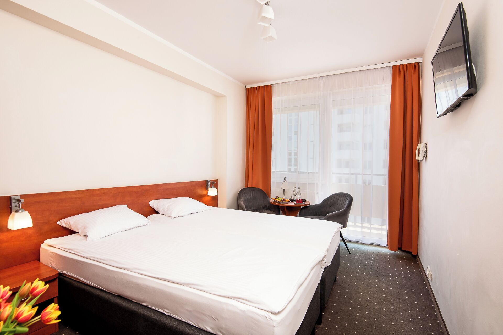 Habitación de hotel con una cama grande, un escritorio con silla, un televisor montado en la pared y una ventana con cortinas.