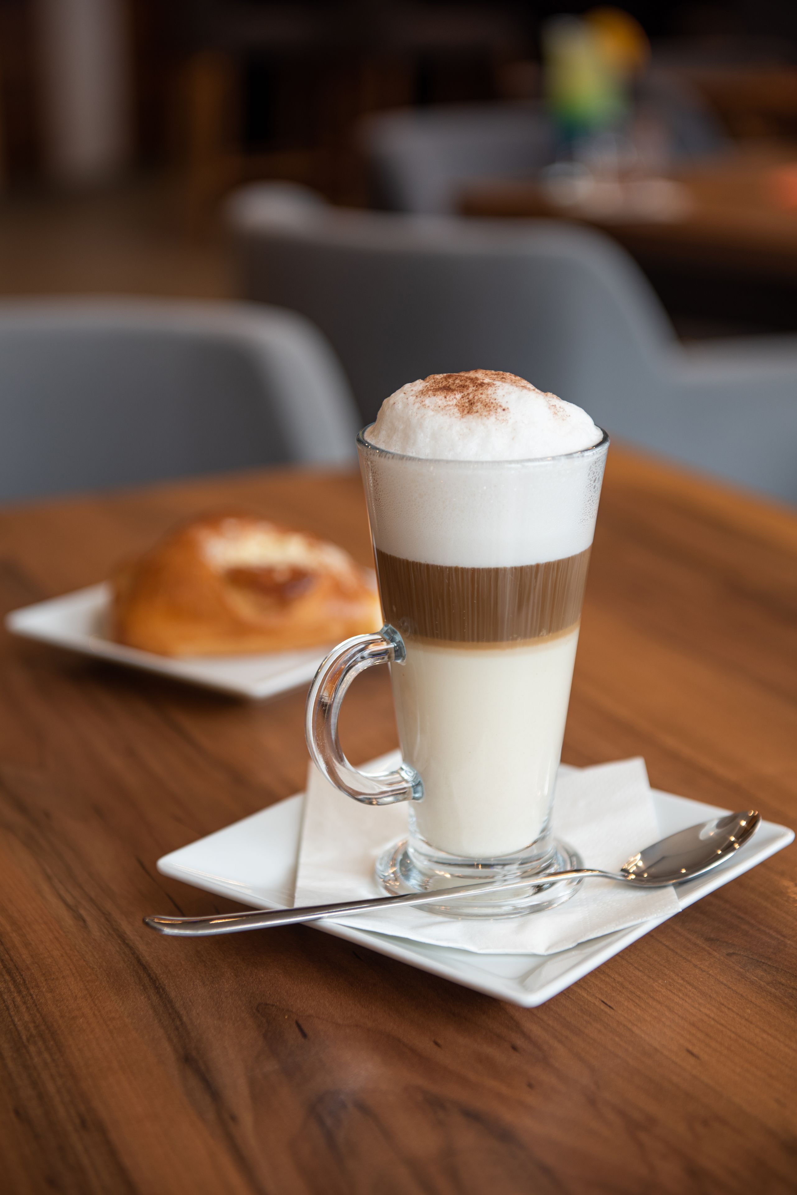 A glass of layered beverage on a wooden table with a pastry in the background.