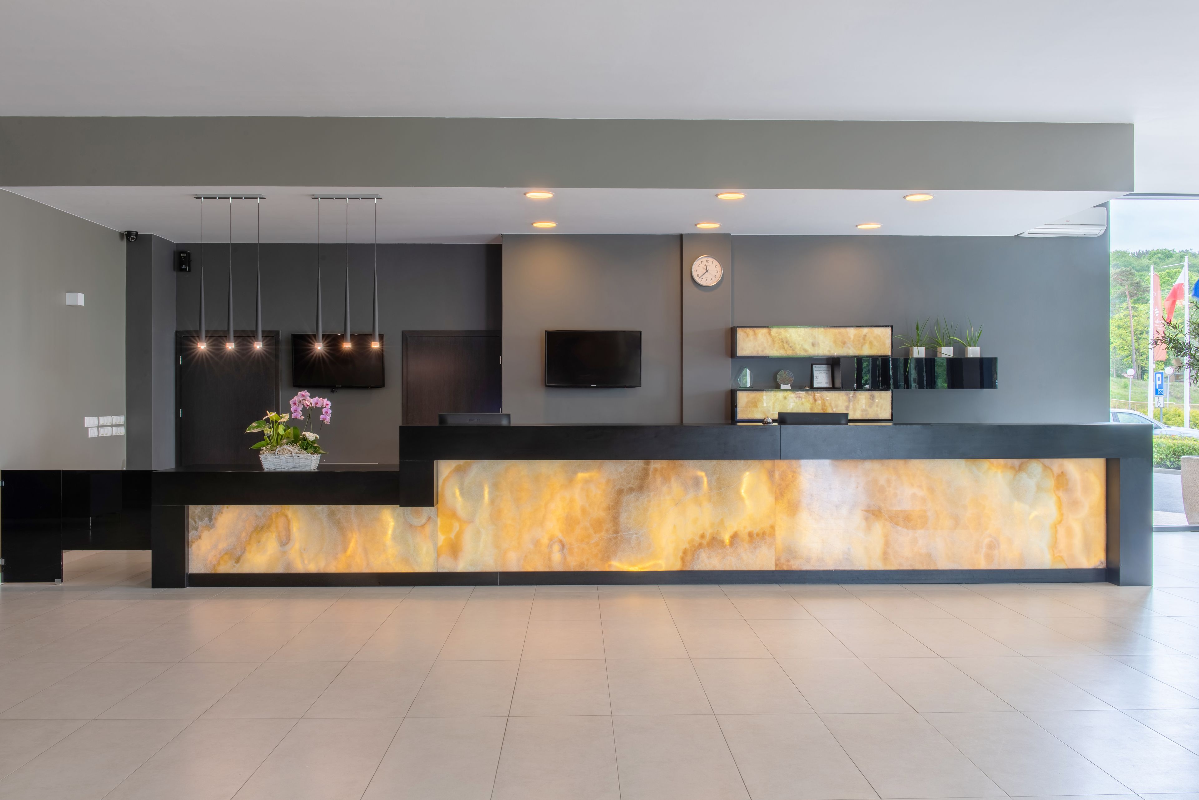 Un espace d’accueil moderne avec un comptoir en marbre, des luminaires suspendus, un téléviseur à écran plat et une horloge murale.