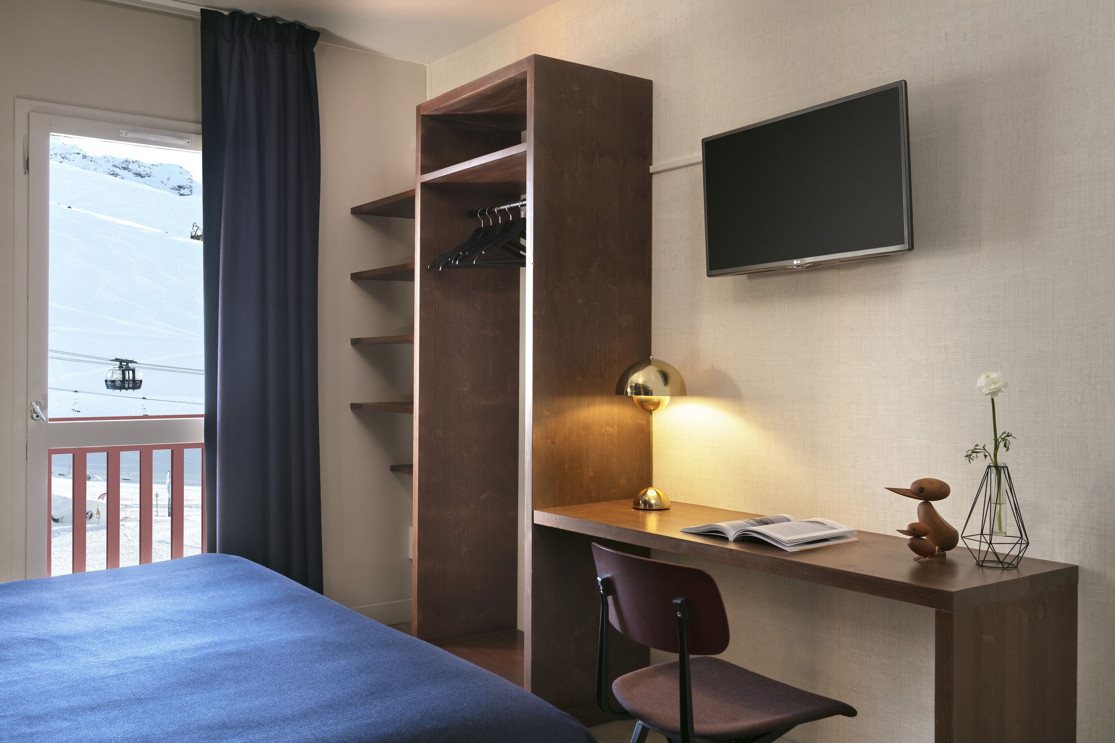 A hotel room with a bed, a desk with a chair, a TV, a window with a view of a snowy mountain, and a wardrobe.