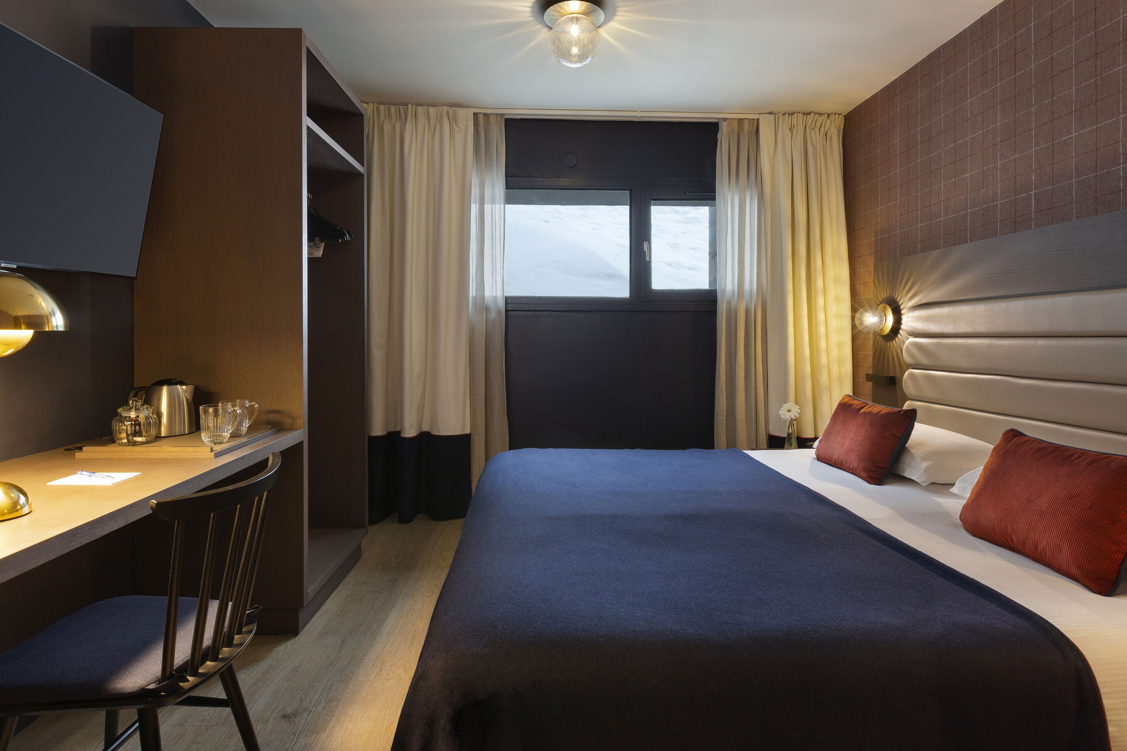 A hotel room with a bed, a desk with a chair, a television, and a window with curtains.