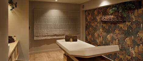 A massage room with a wooden bench, a small table with a towel, and a wall with a patterned tapestry.