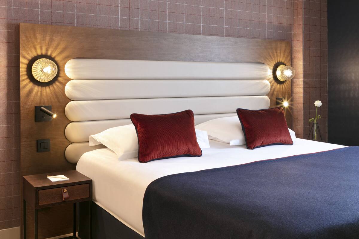 Una habitación de hotel moderna con una cama grande, dos almohadas rojas, una mesita de noche con lámpara y apliques de pared.