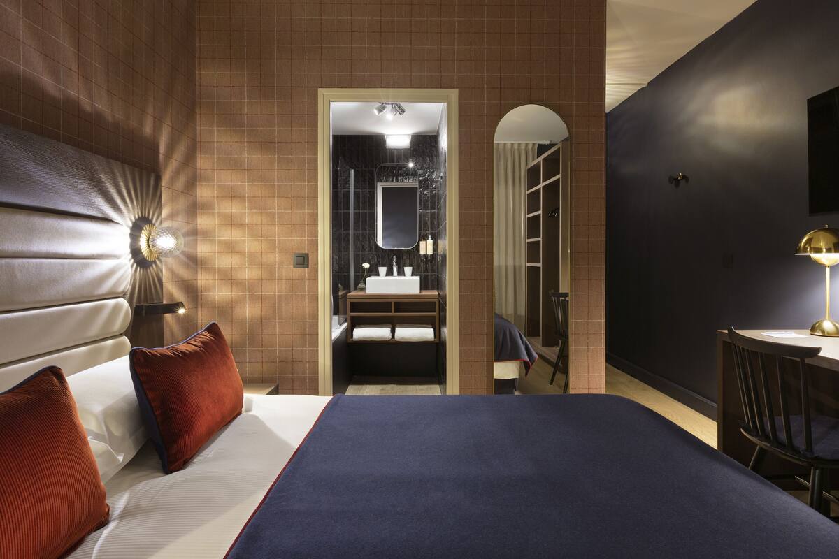 Habitación de hotel con cama, escritorio, silla, espejo y baño visible a través de un arco.