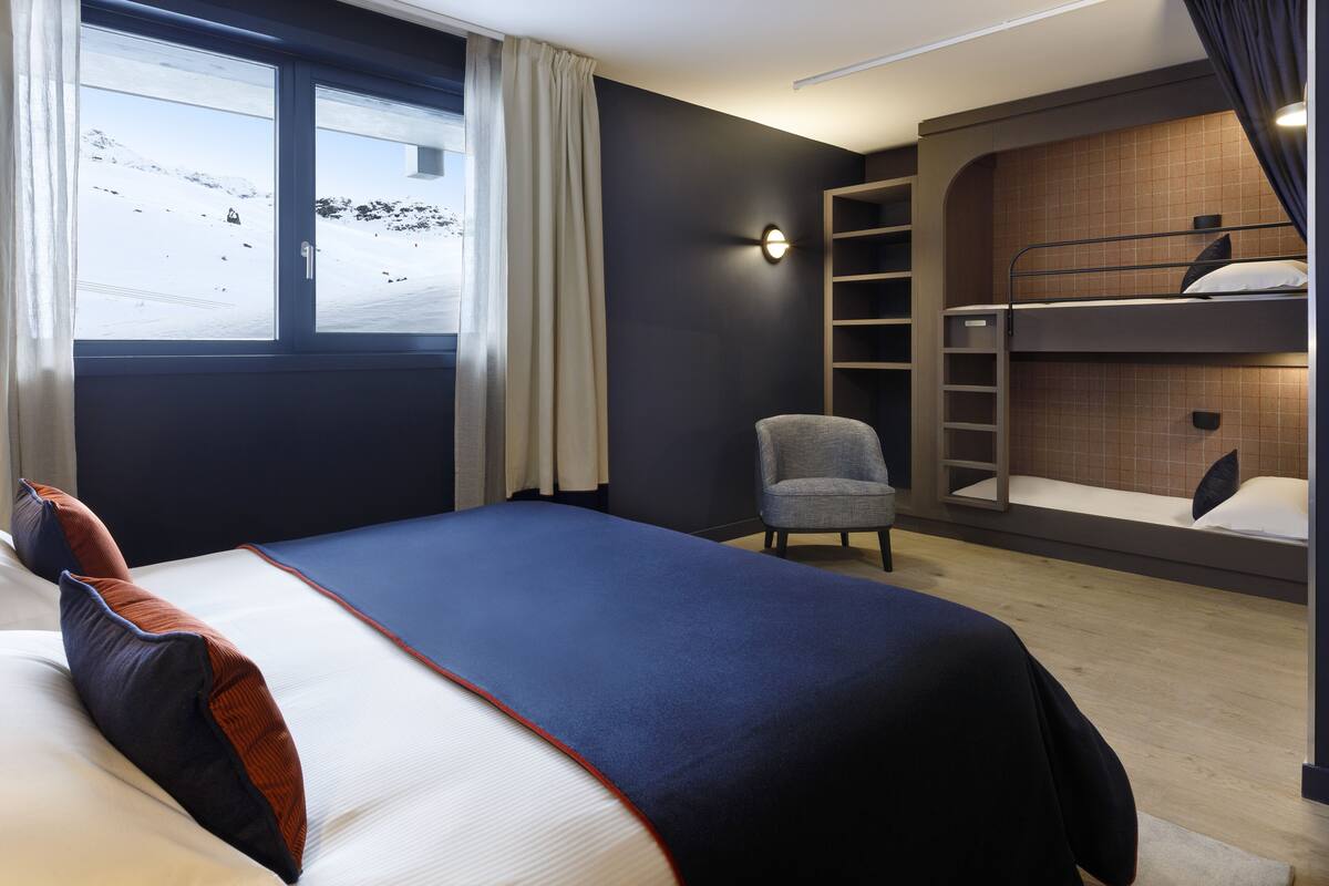 Una habitación de hotel moderna con literas, una cama individual, una silla, una mesita de noche y un ventanal con vistas a un paisaje nevado.