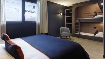 Una habitación de hotel moderna con literas, una cama individual, una silla, una mesita de noche y un ventanal con vistas a un paisaje nevado.